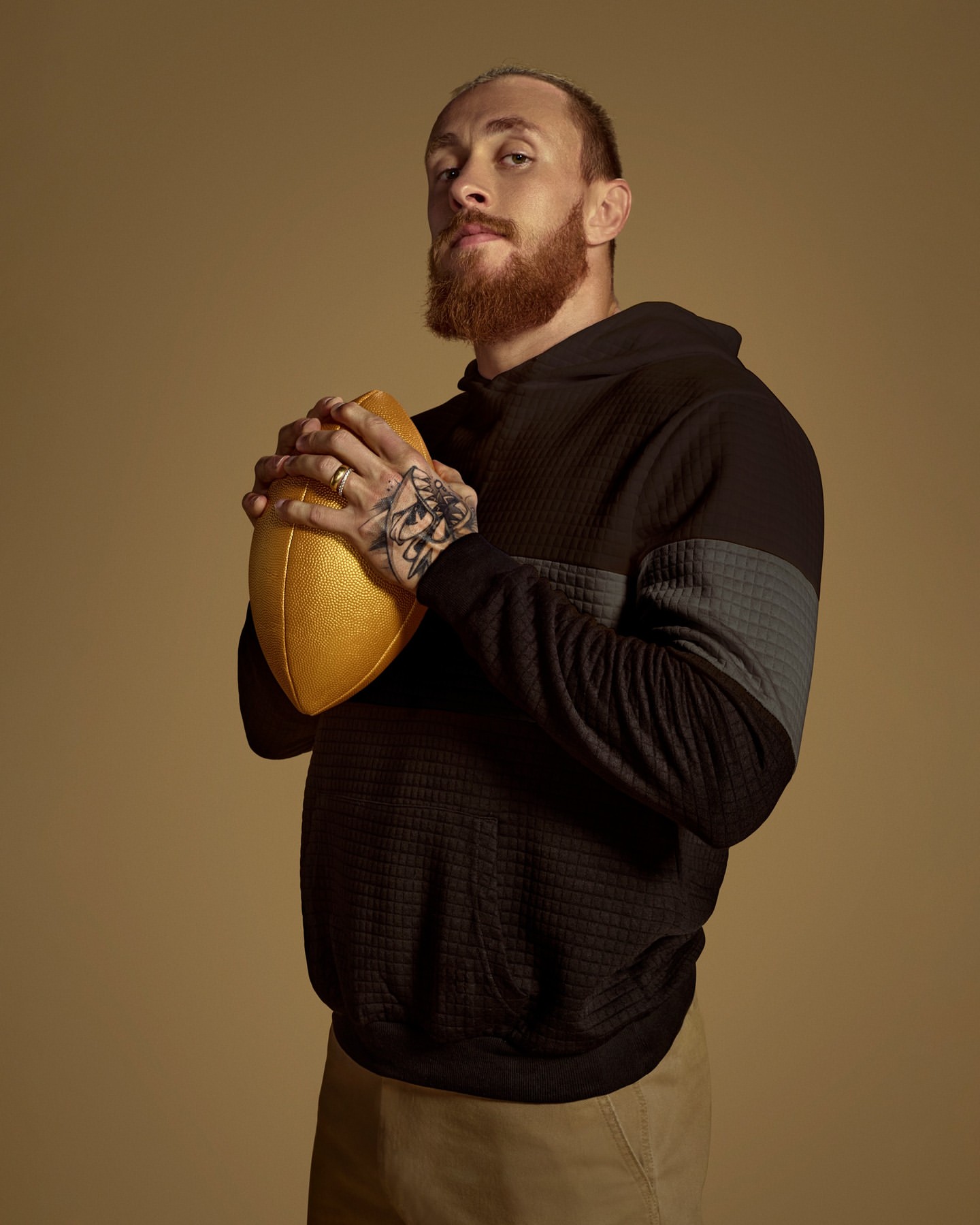 Portrait of a bearded man in a hoodie holding a gold football against a tan background in a photo studio with studio lighting.